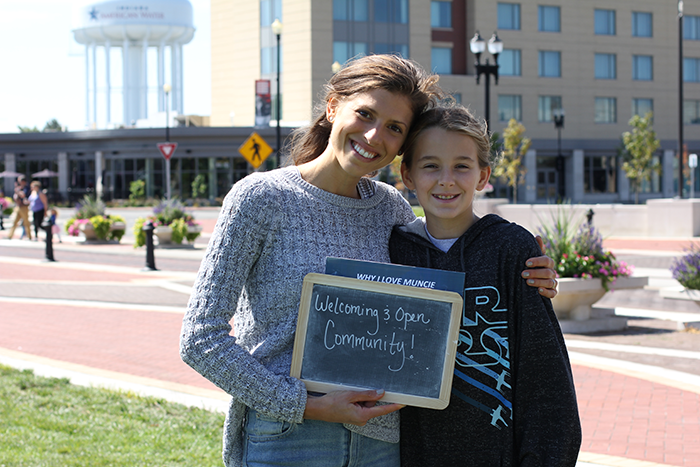 A woman in a gray sweater with her arm around a child in a black hoodie. They are holding a chalkboard on which is written "Welcoming and Open Community!"