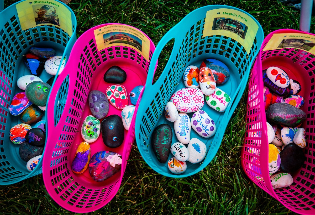 Four colorful baskets, each labeled with a different location, sit in the grass. Each contains many decorated rocks.