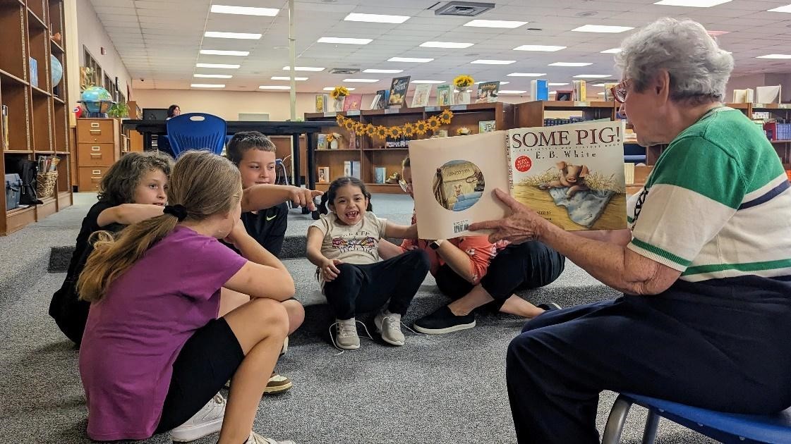 A person in a green and white shirt sits on a low chair reading to children gathered around in an arc around him on the floor