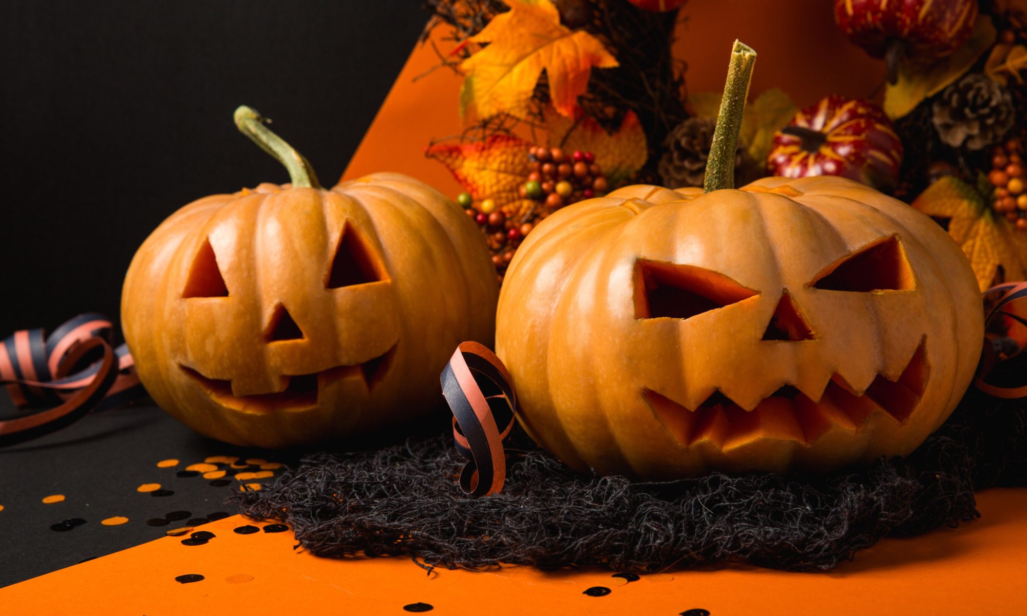 Two jack'o'lanterns sitting on a black and orange table. The back one has a smiley face and one more toward the front has a scary face.