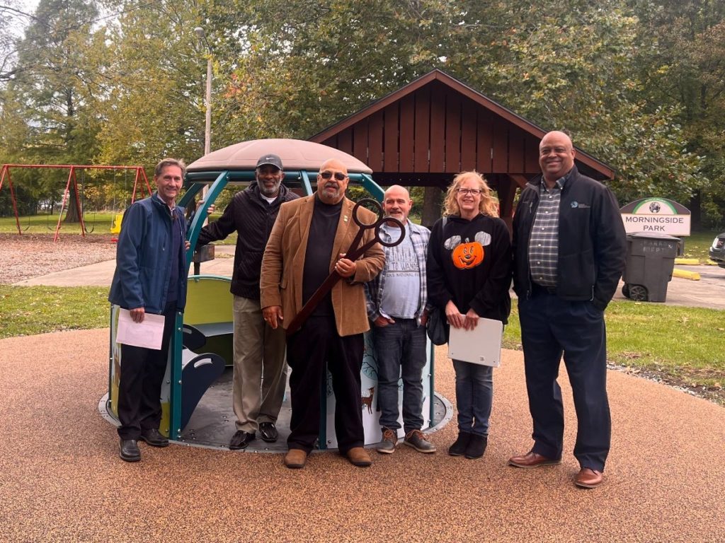 Mayor Ridenour stands with Neighborhood Association members in front of the new We-Go-Round. A man in the center of the group holds a large pair of black, ribbon-cutting scissors. 