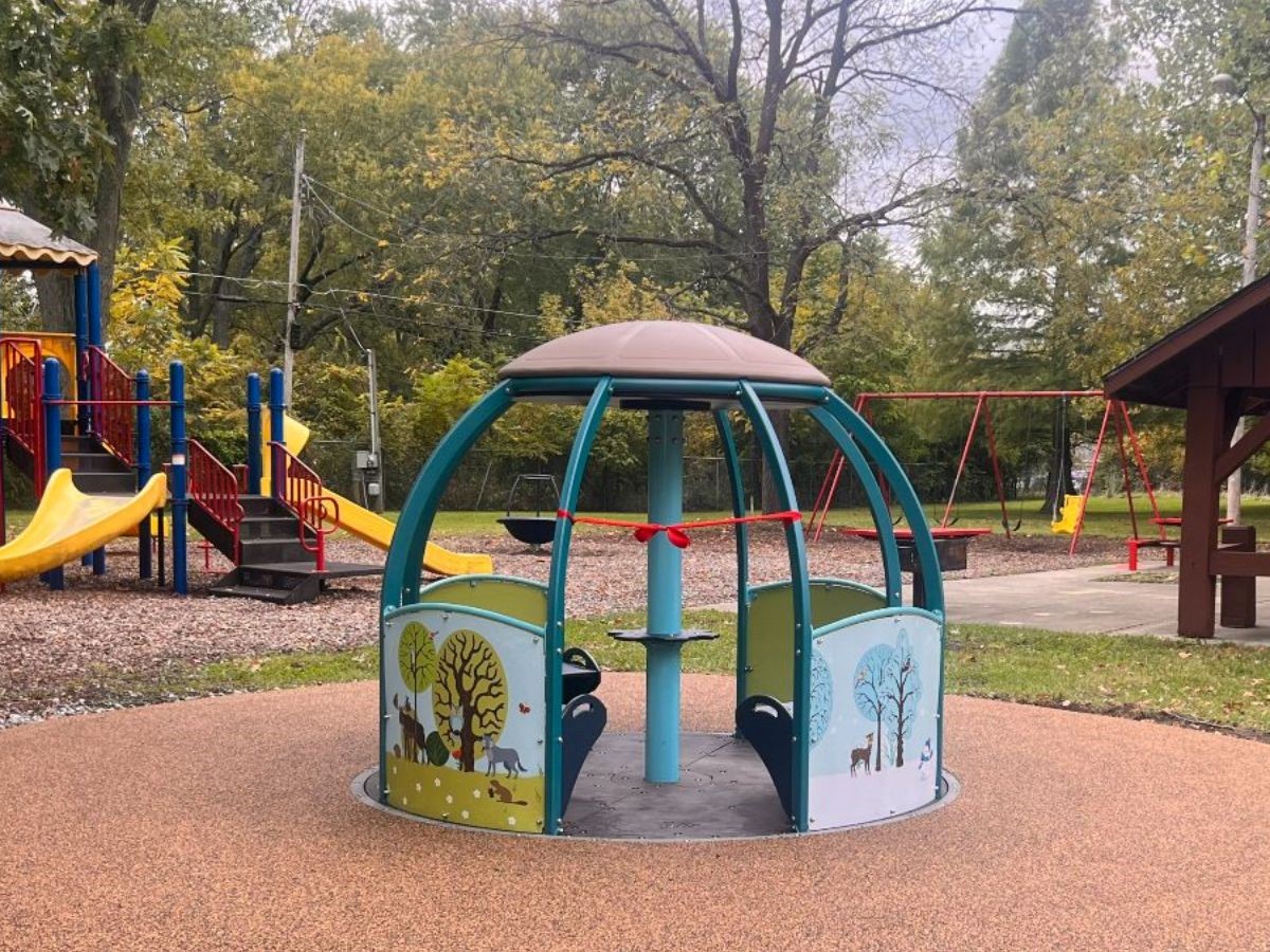 A dome with vertical spokes sits on an attractive slab against the background of the rest of the park. It has seats inside and the backs of the seats are painted with seasonal motifs. Visible on the front are a winter scene with a deer and three snow-covered trees, and a scene with two trees and a variety of animals including a beaver and a wolf facing the center of the panel.