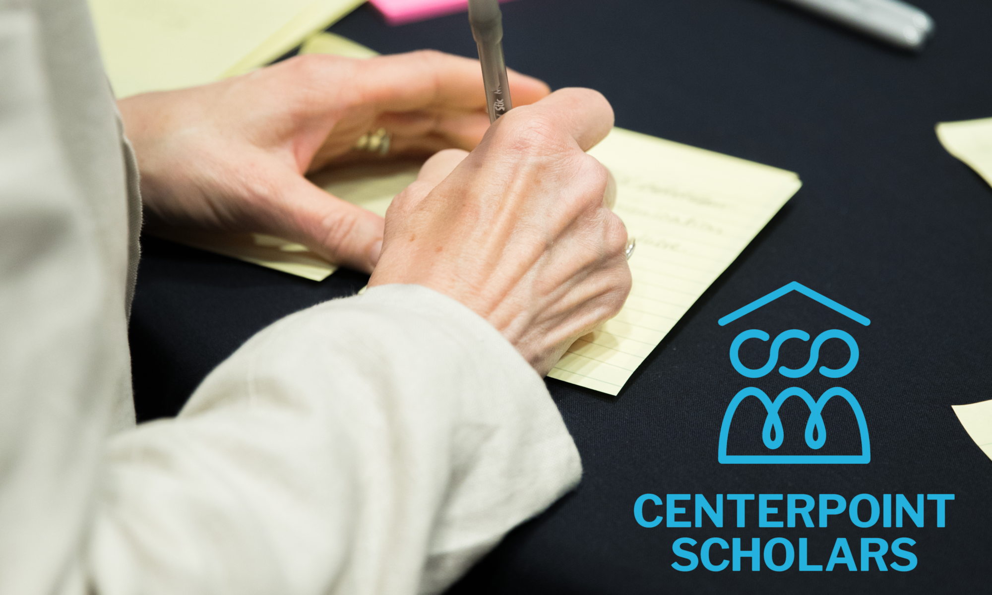 A woman's hands writing on a yellow paper set on a black table cloth. The CenterPoint Scholars logo is set off to her right. There are other yellow papers arranged around the table with headers like "Residential Policies"