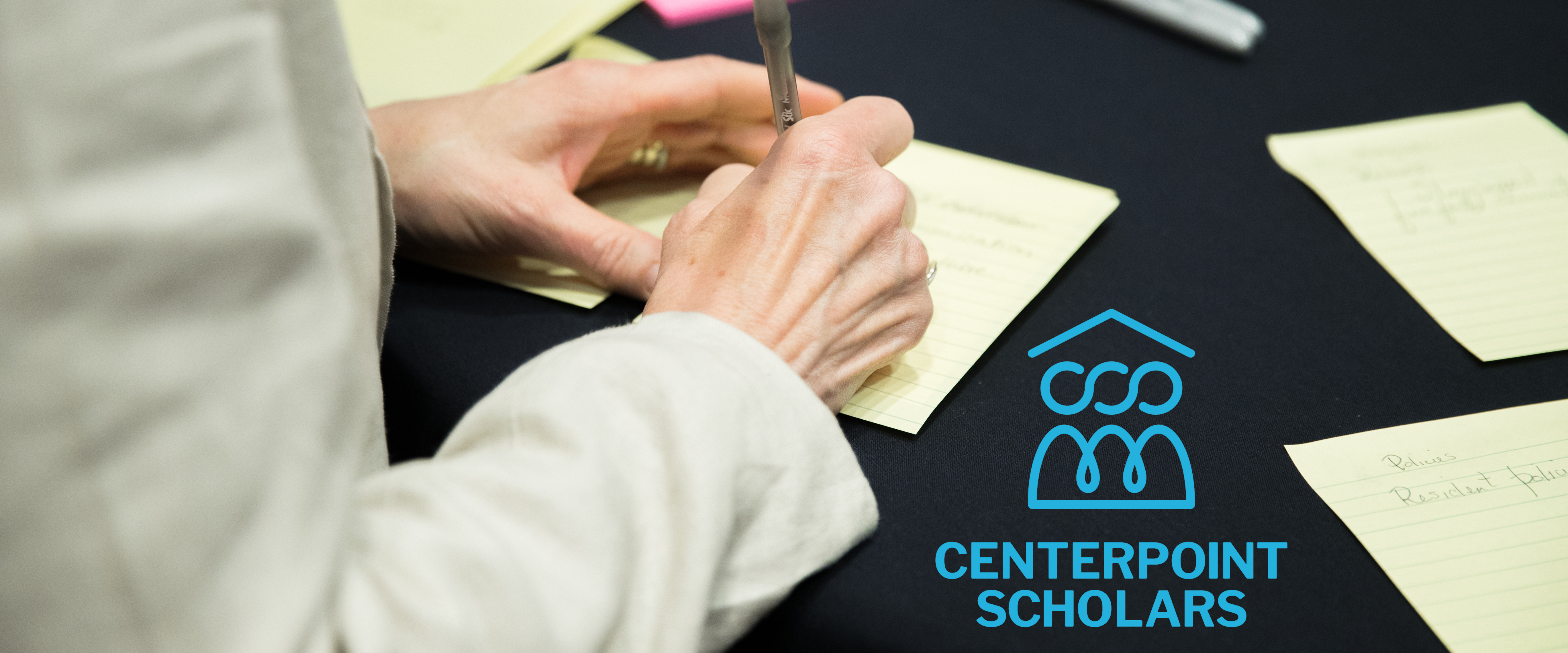 A woman's hands writing on a yellow paper set on a black table cloth. The CenterPoint Scholars logo is set off to her right. There are other yellow papers arranged around the table with headers like "Residential Policies"